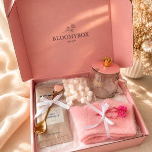 May include: A pink gift box labelled BLOOMYBOX SHOP containing a glass mug with a gold crown lid, a packet of milk chocolate cocoa, a bag of white marshmallows, a gold heart-topped spoon, and pink fuzzy socks tied with a white ribbon.
