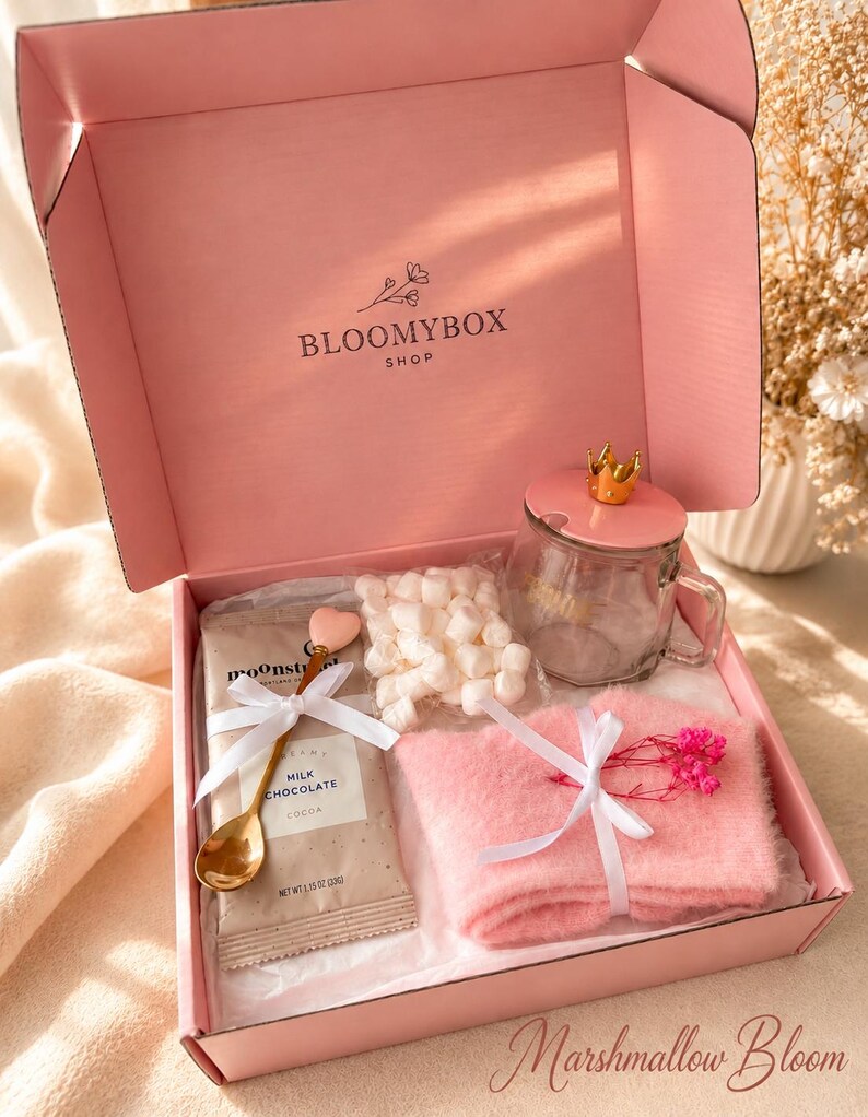 May include: A pink gift box labelled BLOOMYBOX SHOP containing a glass mug with a gold crown lid, a packet of milk chocolate cocoa, a bag of white marshmallows, a gold heart-topped spoon, and pink fuzzy socks tied with a white ribbon.