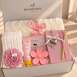 May include: A gift box labelled BLOOMYBOX SHOP containing a cream-coloured knit blanket with a pink ribbon, a pink notebook with a heart keychain, a gold pen, a pink flower-shaped candle, a rose hand cream, and a pink eye mask.