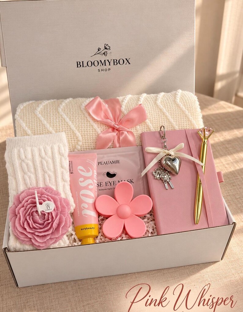 May include: A gift box labelled BLOOMYBOX SHOP containing a cream-coloured knit blanket with a pink ribbon, a pink notebook with a heart keychain, a gold pen, a pink flower-shaped candle, a rose hand cream, and a pink eye mask.