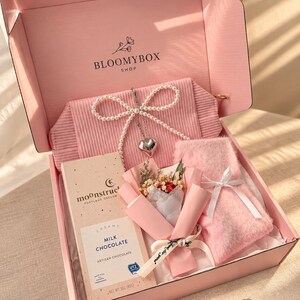 May include: A pink gift box labelled BLOOMYBOX SHOP containing a ribbed pink pouch with a pearl bow and heart charm, an 85g bar of Moonstruck milk chocolate, a small dried flower bouquet, and a pair of fuzzy pink socks tied with a white ribbon.