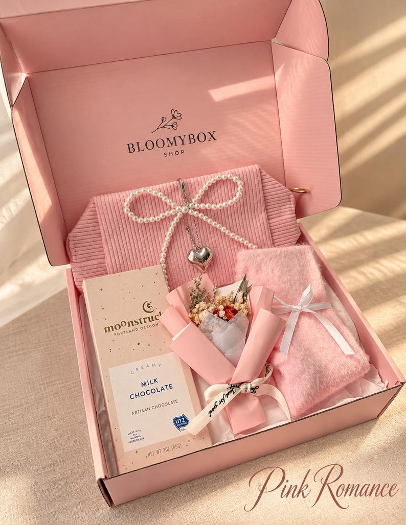 May include: A pink gift box labelled BLOOMYBOX SHOP containing a ribbed pink pouch with a pearl bow and heart charm, an 85g bar of Moonstruck milk chocolate, a small dried flower bouquet, and a pair of fuzzy pink socks tied with a white ribbon.