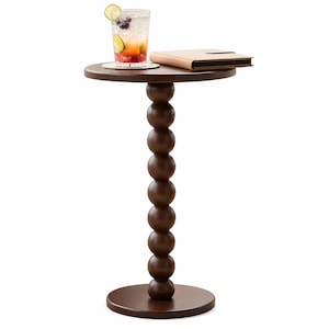 Puede incluir: Una mesa auxiliar de madera marrón oscuro con tablero y base circulares, que presenta una columna central compuesta por esferas de madera apiladas. Un vaso de bebida de frutas con hielo y un cuaderno de cuero beige descansan sobre la mesa.
