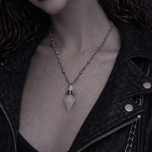 Può includere: Una collana d'argento con un ciondolo a forma di fiala di cristallo. Il ciondolo trasparente e sfaccettato è sospeso a una catena di perline. L'immagine mostra una giacca di pelle nera e trucco scuro.
