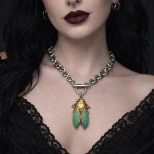 May include: Silver-tone chain necklace with a pendant featuring a detailed insect design. The insect pendant has gold, green, and bronze tones. The necklace is worn against a black lace top.