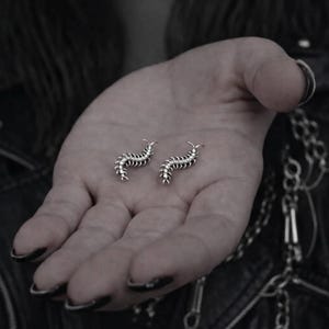 May include: Two silver-colored centipede earrings. Each earring has a segmented body with multiple legs and curved antennae. The earrings are displayed on a person's open palm, with a dark background.