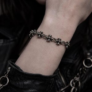 May include: Silver bracelet with cross-shaped charms. The bracelet is worn on a wrist, with a black leather jacket in the background. The crosses are detailed and evenly spaced. The bracelet's design is symmetrical.