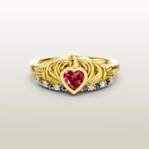 May include: A gold-colored ring with a heart-shaped red gemstone at the center. The ring features a crown design and is accented with small blue and white gemstones. The ring is set against a soft, light background.