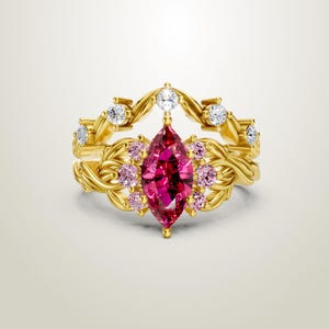 May include: A gold-colored ring set featuring a marquise-cut, deep pink gemstone as the focal point. The ring is adorned with smaller pink and clear gemstones, and has a detailed, vine-like design.