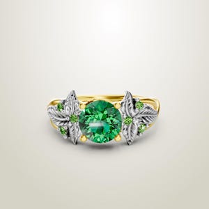 May include: A gold ring with a large, round, emerald-green gemstone at the center. The ring features silver-colored leaf designs on either side of the central stone, accented with smaller green gemstones. The band is gold-colored.