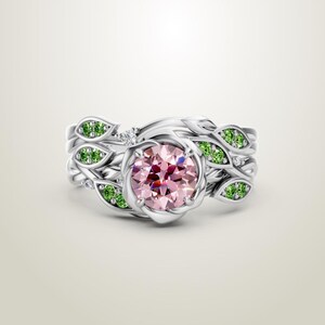 May include: A silver ring set featuring a central pink gemstone surrounded by a floral design. The ring is accented with green gemstones and small white stones, creating a nature-inspired aesthetic. The band has a leaf-like pattern.