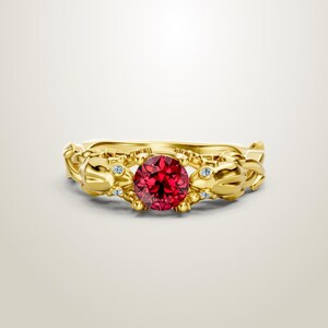 May include: A gold ring with a large, round, red gemstone at the center. The ring has an ornate design with smaller blue gemstones and decorative elements. The ring is set against a white background.