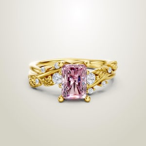 May include: A gold ring set featuring a pink rectangular gemstone as the centerpiece, flanked by smaller white gemstones. The band has a textured, vine-like design, creating an elegant and nature-inspired aesthetic. A matching band sits beneath the main ring.