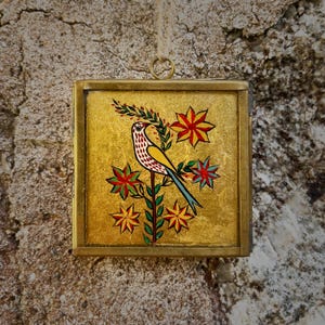 Puede incluir: Obra de arte enmarcada cuadrada con un pájaro posado en una rama floreciente. El pájaro tiene un cuerpo amarillo, marcas rojas y blancas y una cola azul. El fondo es dorado y las flores son rojas y verdes.