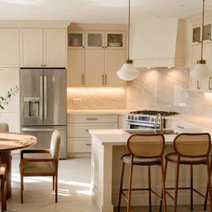 May include: A kitchen interior featuring cream-colored cabinetry, a stainless steel refrigerator, and a marble backsplash. A central island includes a sink and two wooden bar stools with woven cane backrests. Two white pendant lights hang above.