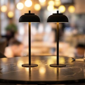 May include: Two black table lamps with a minimalist design. Each lamp has a rounded shade and a slender stem, resting on a circular base. The lamps are positioned on a dark, patterned table, with a blurred background of a restaurant setting.