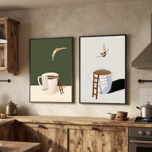 Può includere: Due stampe artistiche incorniciate raffigurano una persona che si tuffa in una tazza di caffè. Una stampa ha uno sfondo verde, l'altra uno sfondo crema. Ogni tazza ha una scala e un asciugamano. Le stampe sono esposte in una cucina con mobili in legno.