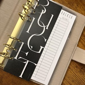 May include: A beige planner with gold-colored rings, holding a black and white "BUDGET" divider and a "TRACKER BALANCE" sheet. The planner is open, revealing the interior pages and the ring mechanism.