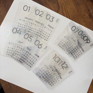 May include: A set of clear, rectangular calendars for the year 2026. Each calendar displays a different month, with the month and year printed in a modern font. The calendars are arranged on a white surface.
