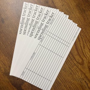 May include: A stack of white spending tracker sheets with black text and ruled lines for tracking expenses. The sheets are labeled with "spending tracker", "DATE", "CATEGORY", "AMOUNT", and "BALANCE". The phrase "every dollar gently counted" is printed at the bottom.