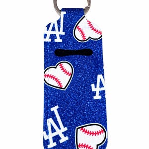May include: A blue fabric keychain holder featuring a repeating pattern of white LA logos and white heart-shaped baseballs with red stitching. The rectangular holder has a rounded bottom and a silver metal split ring attached at the top.