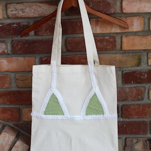 May include: Cream-colored tote bag with white straps and a decorative bikini top design. The bikini top features green and white gingham triangles with white lace trim. The bag hangs on a wooden hanger against a brick wall.