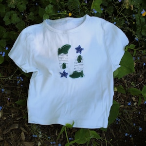 May include: White t-shirt with a crew neck and short sleeves. The shirt features a design of two green and white boots with blue star accents. The shirt is laying on a bed of green foliage and brown mulch.