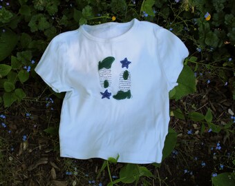 Peut inclure: T-shirt blanc à col rond et manches courtes. Le t-shirt présente un motif de deux bottes vertes et blanches avec des étoiles bleues. Le t-shirt est posé sur un lit de feuillage vert et de paillis brun.
