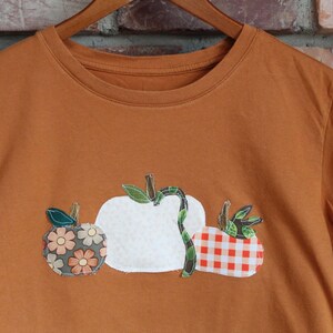 Peut inclure: T-shirt orange br&ucirc;l&eacute; avec trois citrouilles appliqu&eacute;es. Une citrouille est blanche, une autre &agrave; motifs floraux et la troisi&egrave;me &agrave; carreaux rouges et blancs. Le t-shirt est suspendu &agrave; un cintre en bois.