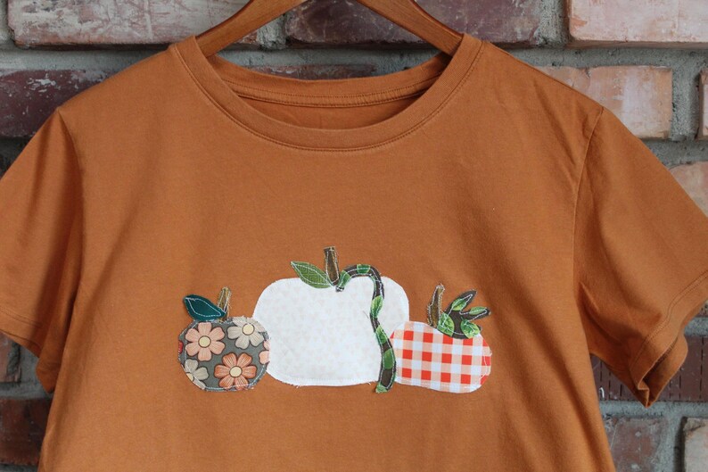 Peut inclure: T-shirt orange br&ucirc;l&eacute; avec trois citrouilles appliqu&eacute;es. Une citrouille est blanche, une autre &agrave; motifs floraux et la troisi&egrave;me &agrave; carreaux rouges et blancs. Le t-shirt est suspendu &agrave; un cintre en bois.