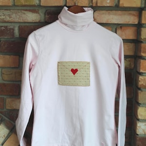 May include: A light pink turtleneck long-sleeve shirt. The shirt features a rectangular patch with a red heart and a dotted pattern. The shirt is hanging on a wooden hanger against a brick wall.