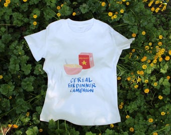 Peut inclure: T-shirt blanc avec un dessin d'un bol de céréales rose, une boîte de céréales rouge et le texte bleu "Cereal for Dinner Campaign". Le t-shirt est posé sur un fond de feuillage vert et de fleurs jaunes.