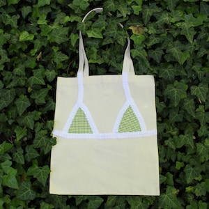 May include: A pale yellow tote bag with white straps and a white trim design. The bag has two green and white checked triangle shapes, resembling a bikini top. The bag is made of a soft material and is suitable for carrying items.