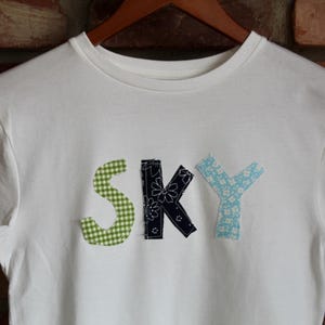 May include: White t-shirt with the word "SKY" appliqu&eacute;d on the front. The letter "S" is green gingham, the "K" is navy with white floral accents, and the "Y" is light blue with a floral pattern.