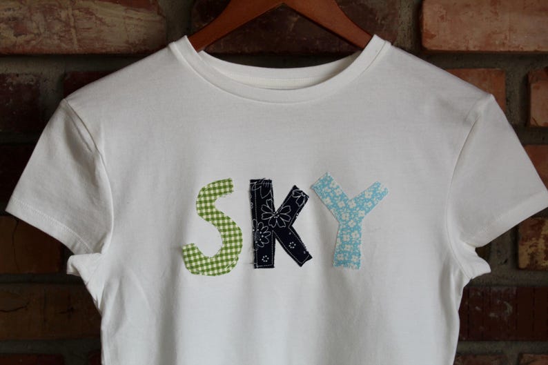 May include: White t-shirt with the word "SKY" appliqu&eacute;d on the front. The letter "S" is green gingham, the "K" is navy with white floral accents, and the "Y" is light blue with a floral pattern.