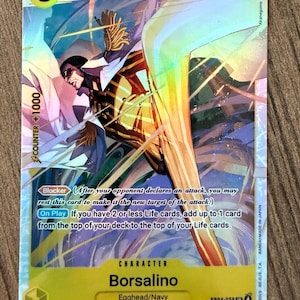 Puede incluir: Una tarjeta coleccionable con el personaje Borsalino de Egghead/Navy. La tarjeta tiene un esquema de color amarillo y azul con el número 5 y el texto "COUNTER +1000". La tarjeta también tiene el texto "Blocker" y "On Play".