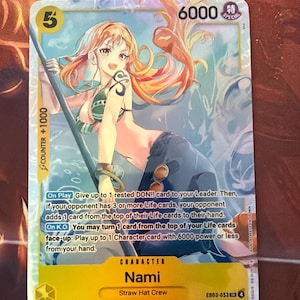 May include: A trading card featuring Nami from the Straw Hat Crew. The card has a yellow border with the number 5 and 6000. The illustration shows Nami with orange hair, a tattoo, and a green top. Text on the card describes gameplay.