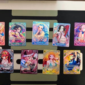 May include: A collection of colorful trading cards featuring anime-style illustrations of female characters. Each card has a unique design, with varying color schemes and character poses. The cards are arranged on a dark surface.