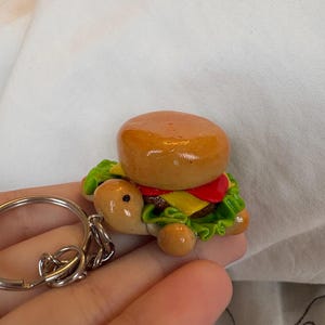 May include: A novelty keychain featuring a turtle-shaped burger. The burger has a golden-brown bun, red tomato, yellow cheese, and green lettuce. The turtle's body is tan with small, round feet. A silver keyring is attached.