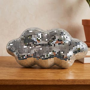 May include: A cloud-shaped decorative object covered in small, reflective mirrored tiles. The silver-colored cloud sits on a wooden surface, reflecting light. The decorative piece is a unique home decor item.