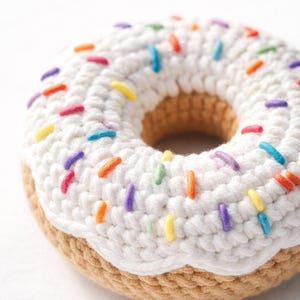 Puede incluir: Un donut tejido a ganchillo con glaseado blanco y chispas de colores. El donut tiene una base marrón y una capa de glaseado blanco con chispas de varios colores, incluyendo rojo, naranja, amarillo, verde, azul y morado.