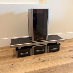 May include: A silver and black XTOOL laser machine with a large rectangular top unit and three drawers below. The machine sits on a wooden floor against a white wall.