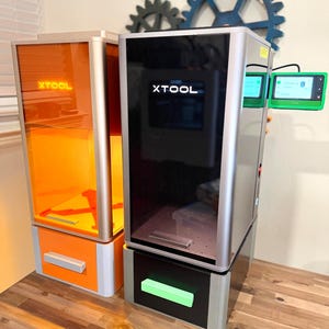 May include: Two XTOOL laser engraving machines. One has an orange interior and the other has a black interior. Both have the XTOOL logo on the front. The machines are on a wooden surface.