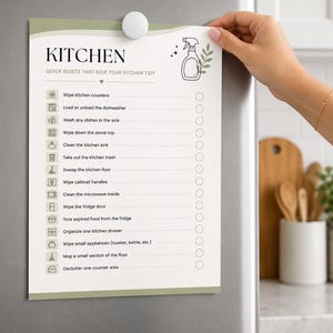 Op de afbeelding: Een witte papieren checklist met de titel KITCHEN QUICK RESETS THAT KEEP YOUR KITCHEN TIDY, een icoon van een spuitfles en een lijst met schoonmaaktaken zoals aanrechten afnemen, vaatwasser inruimen en gootsteen reinigen met vakjes.