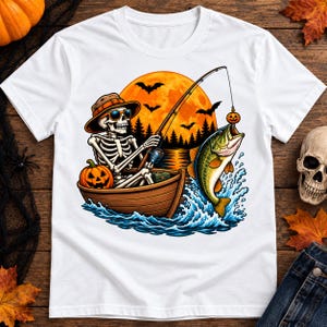 Op de afbeelding: Een wit t-shirt met een afbeelding van een skelet met een hoed en zonnebril, zittend in een houten boot op het water. Het skelet vist naar een grote groene vis met een pompoenvormig kunstaas tegen een grote oranje maan en vleermuizen.