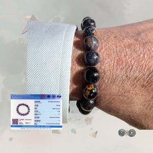 Könnte beinhalten: Ein Armband mit dunkelblauen und schwarzen kugelförmigen Perlen mit goldenen Einschlüssen. Das Armband wird am Handgelenk getragen, mit einer weißen Hemdmanschette im Hintergrund. Die Perlen haben einen Durchmesser von ca. 2 cm. Schmuck.
