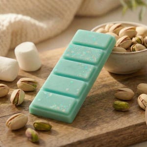 Puede incluir: Una barra de cera perfumada turquesa con cinco secciones, presentada sobre una tabla de madera con pistachos y malvaviscos. Un pequeño cuenco de pistachos y una manta de punto beige están en el fondo. El logotipo "The Scent Nest Home fragrance" es visible.