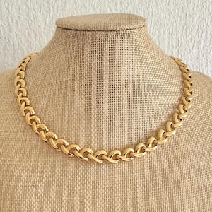 May include: A gold-coloured chain necklace with a repeating link design. The necklace is displayed on a tan mannequin bust against a textured white background. The necklace is a choker style.
