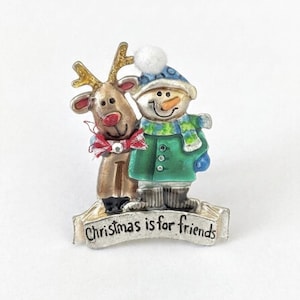 May include: A metal brooch featuring a brown reindeer with a red nose and a red bow, standing next to a snowman wearing a blue hat with a white pom-pom, a green coat, and a green scarf. A silver banner at the bottom reads, Christmas is for friends.