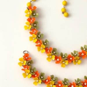 May include: A necklace and bracelet set featuring a series of small, five-petaled flowers in yellow, orange, and olive green. Each flower is accented with a small, clear rhinestone at the center. The necklace includes a strand of yellow beads.